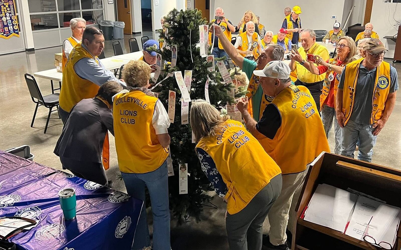 Colleyville Lions in gold club vests gather around a Christmas tree hung with paper gift tags at a club meeting, while other members smile and take photos